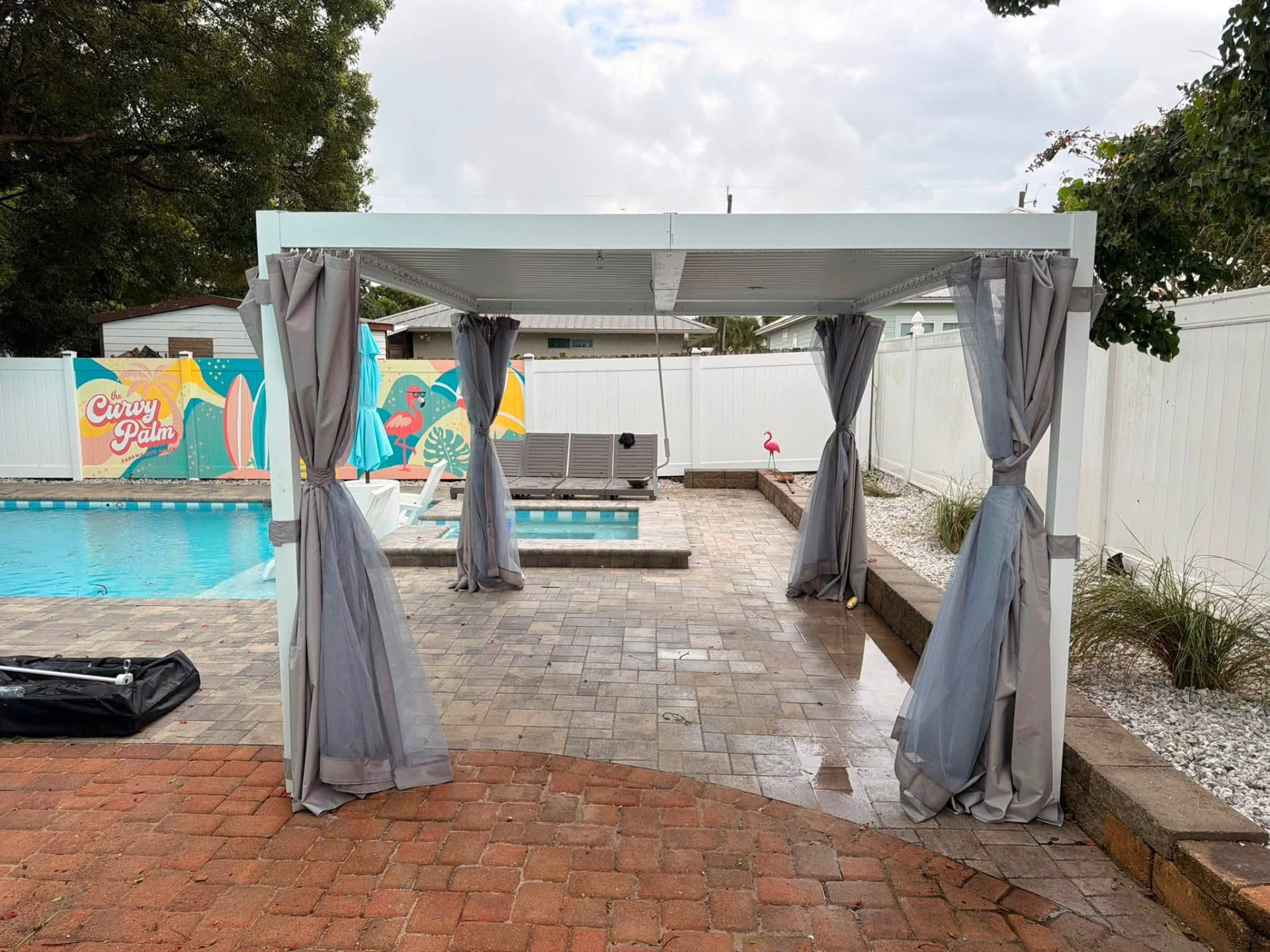 White louvered pool cabana with gray drapes on a paver patio